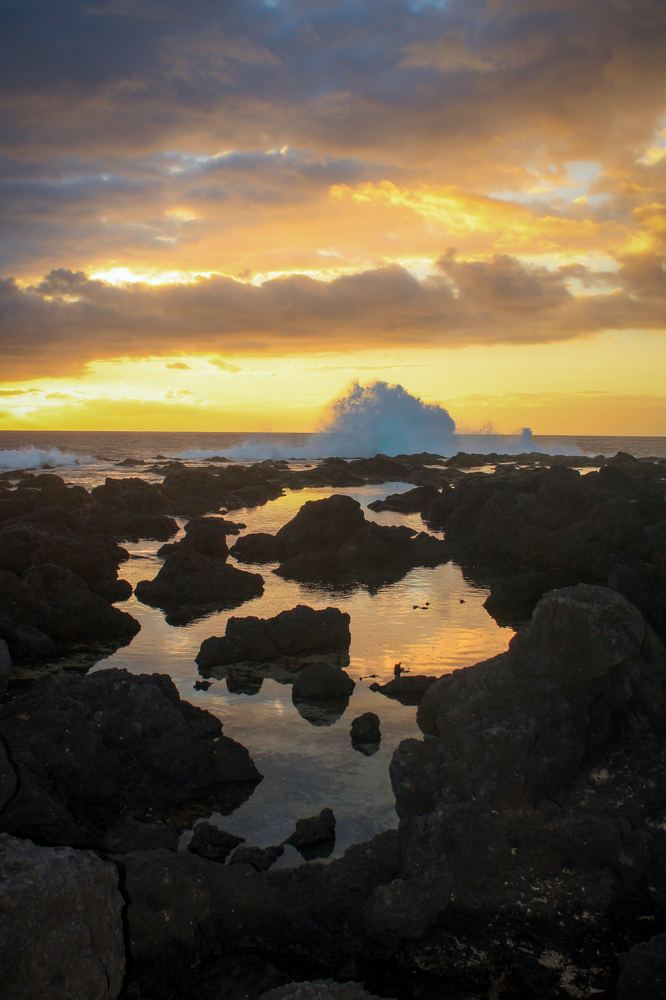 Coucher de soleil sur un bassin d'eau de mer entre les rochers de lave, une vague se brise au loin